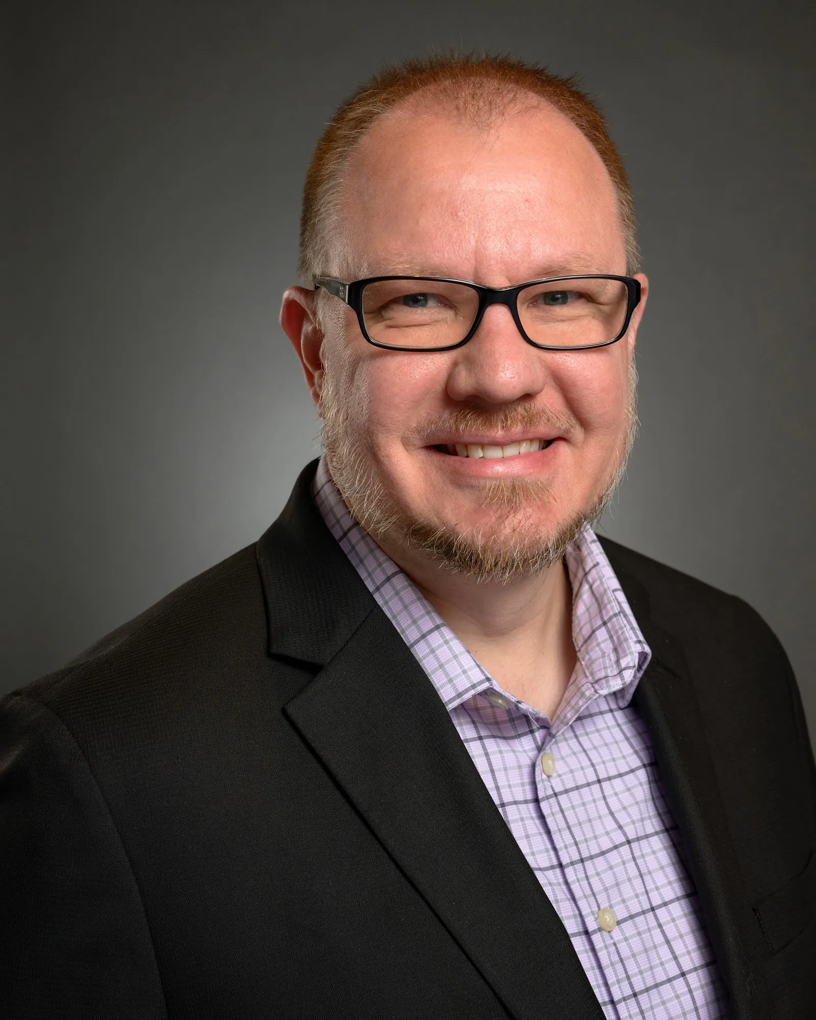 Team headshot, man in glasses and a dark blazer on a grey backdrop