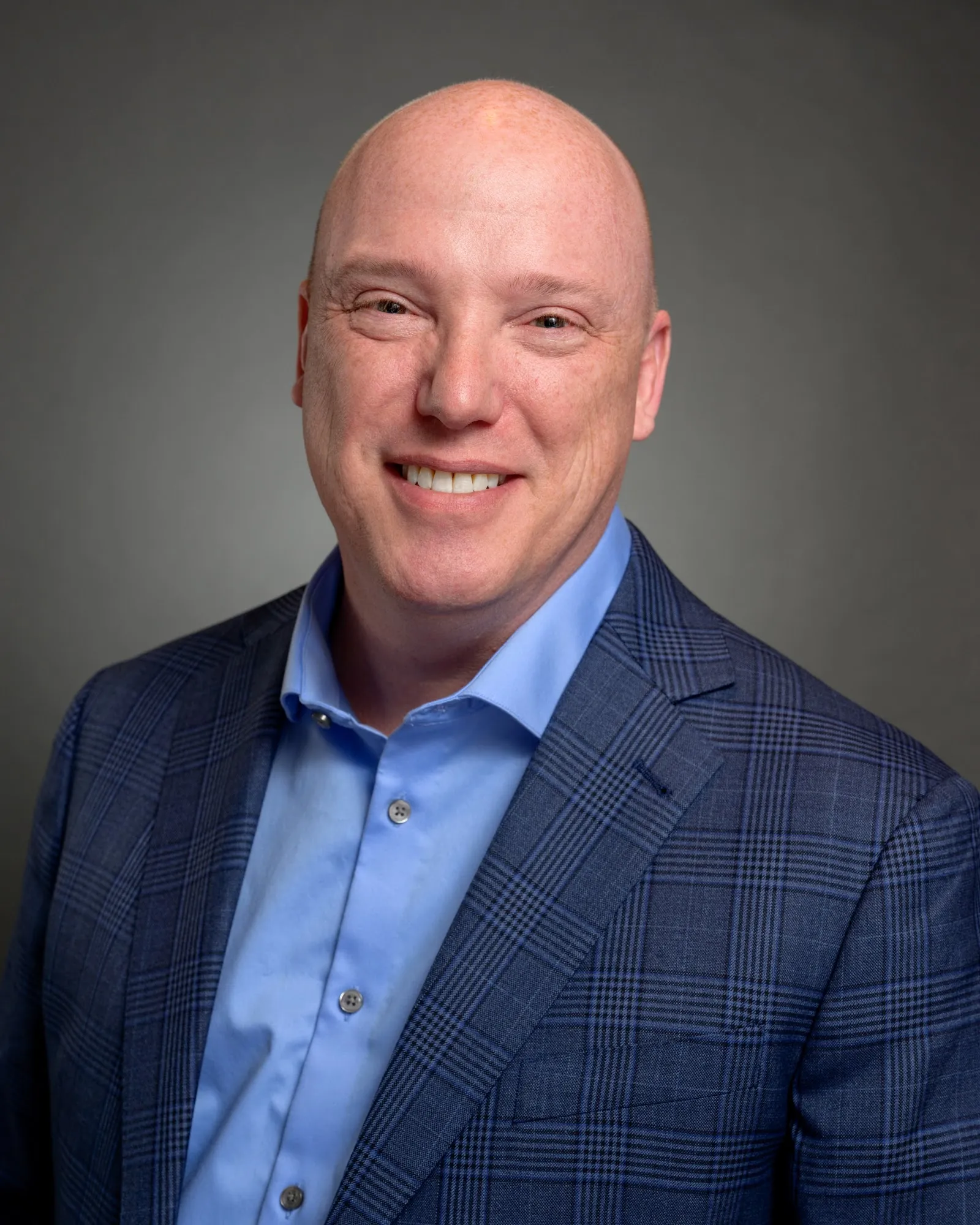 Team headshot, smiling man in a blue plaid blazer on a grey backdrop