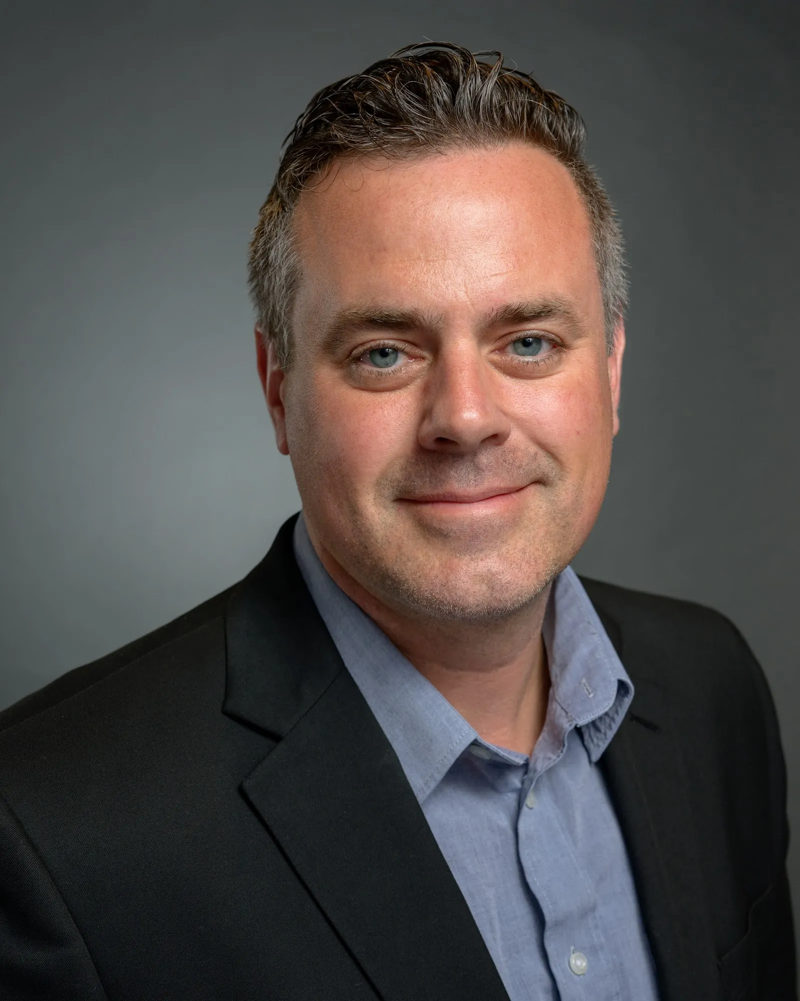 Team headshot, man in a dark blazer and blue shirt on a grey backdrop